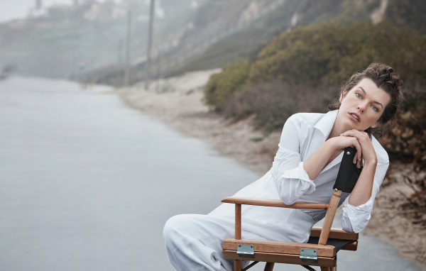 Актриса Милла Йовович в белом платье сидит на стуле