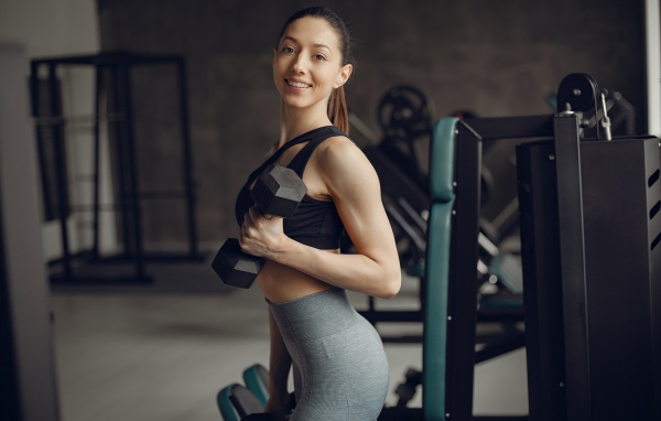 Улыбающаяся спортивная стройная девушка в спортзале с гантелями