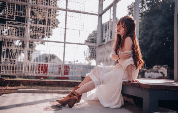 Красивая девушка азиатка в белом платье сидит на лавочке