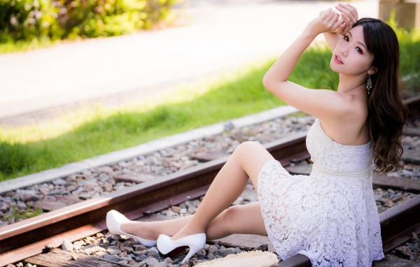 Красивая девушка азиатка в белом платье сидит на рельсах 