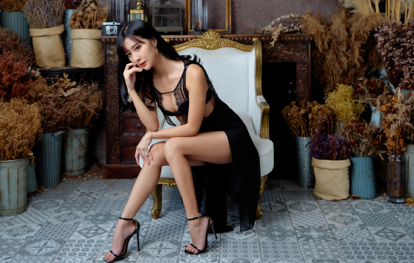 Красивая девушка азиатка в черном платье сидит в белом кресле 