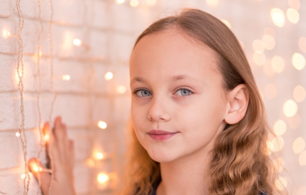 Красивая голубоглазая девочка стоит у стены с гирляндой