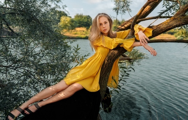Красивая девушка в желтом платье лежит на дереве у воды
