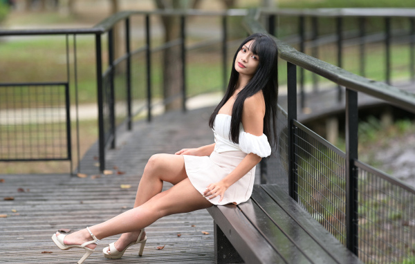 Красивая длинноволосая азиатка в белом платье сидит на лавке