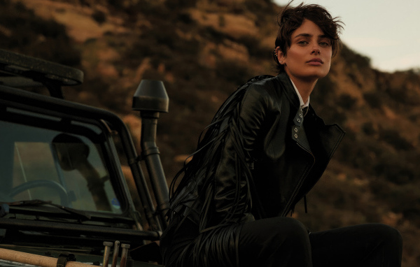 Модель Тейлор Хилл в черном пальто сидит на машине