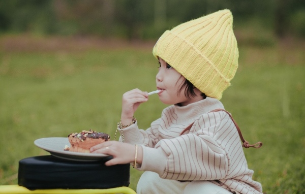 Маленькая девочка в желтой шапке ест пирожное