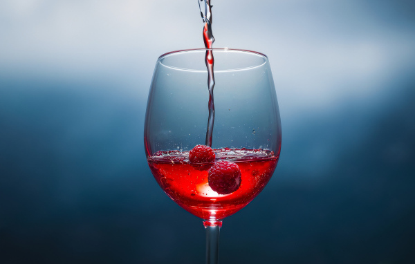 Вино наливают в бокал с ягодами малины на сером фоне