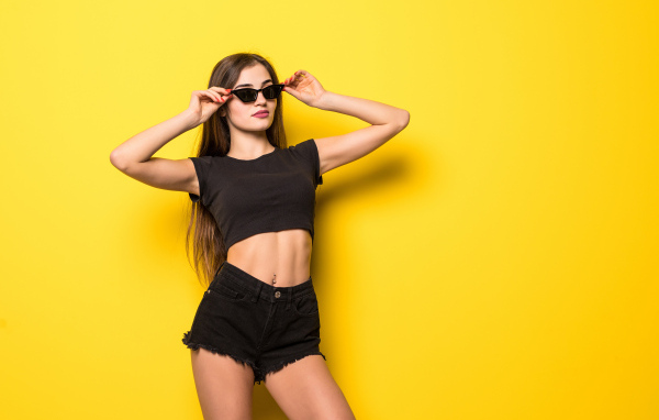 Красивая девушка в черном наряде и очках стоит на желтом фоне