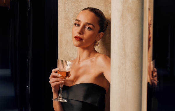 Актриса Эмилия Кларк с бокалом стоит у стены
