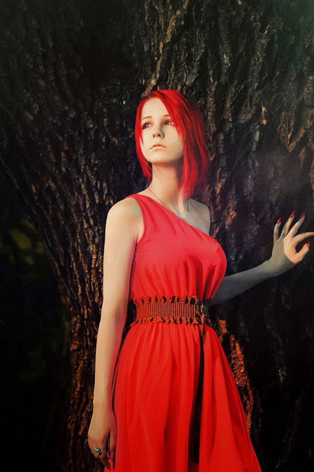 Рыжая девушка в красном платье стоит у дерева