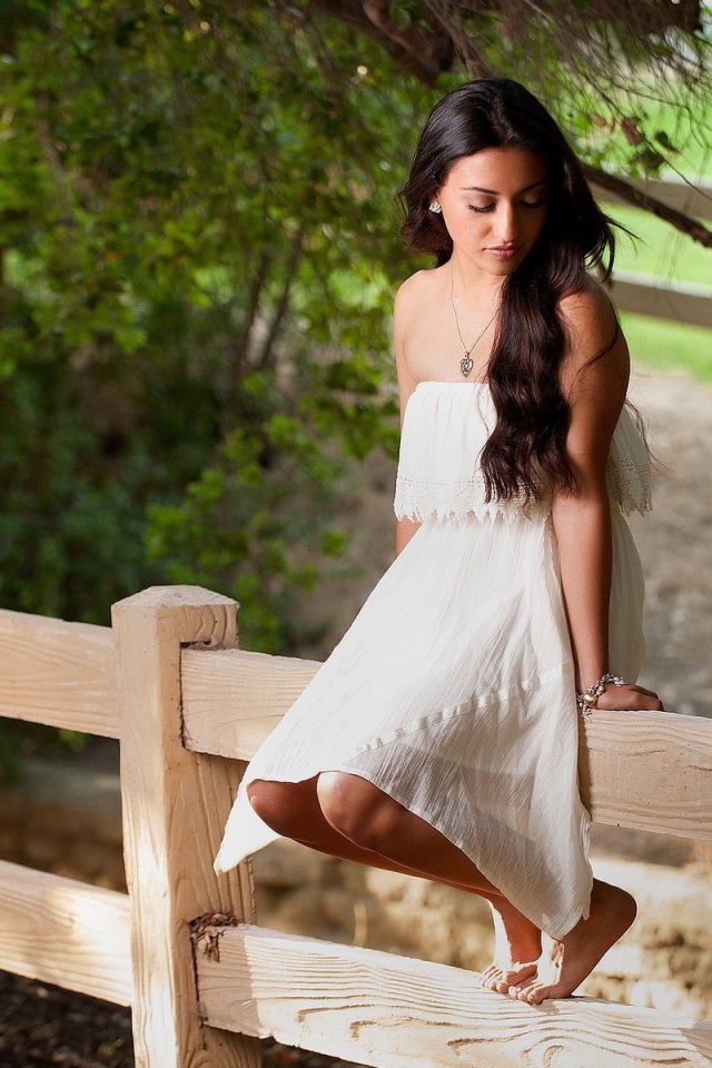 Брюнетка в белом платье сидит на заборе