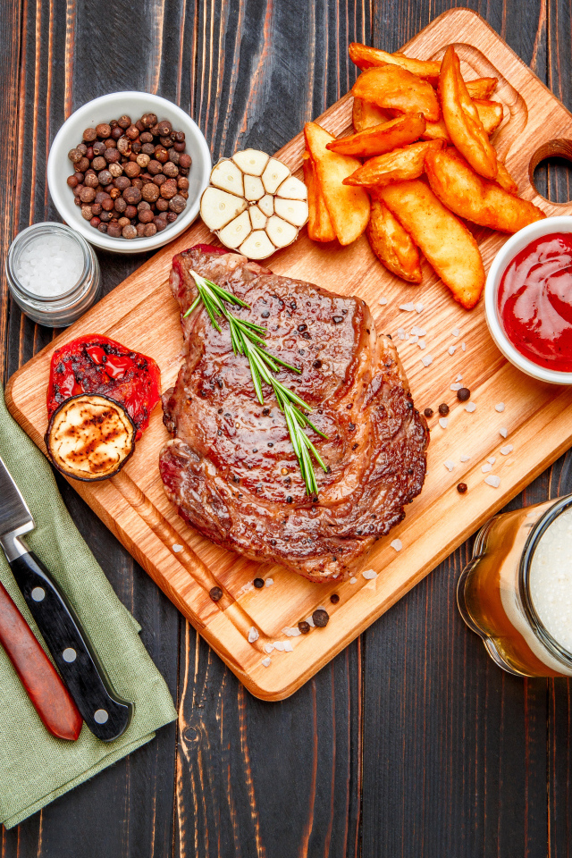 Сочный кусок мяса с картофелем фри, специями и бокалом пива на толе