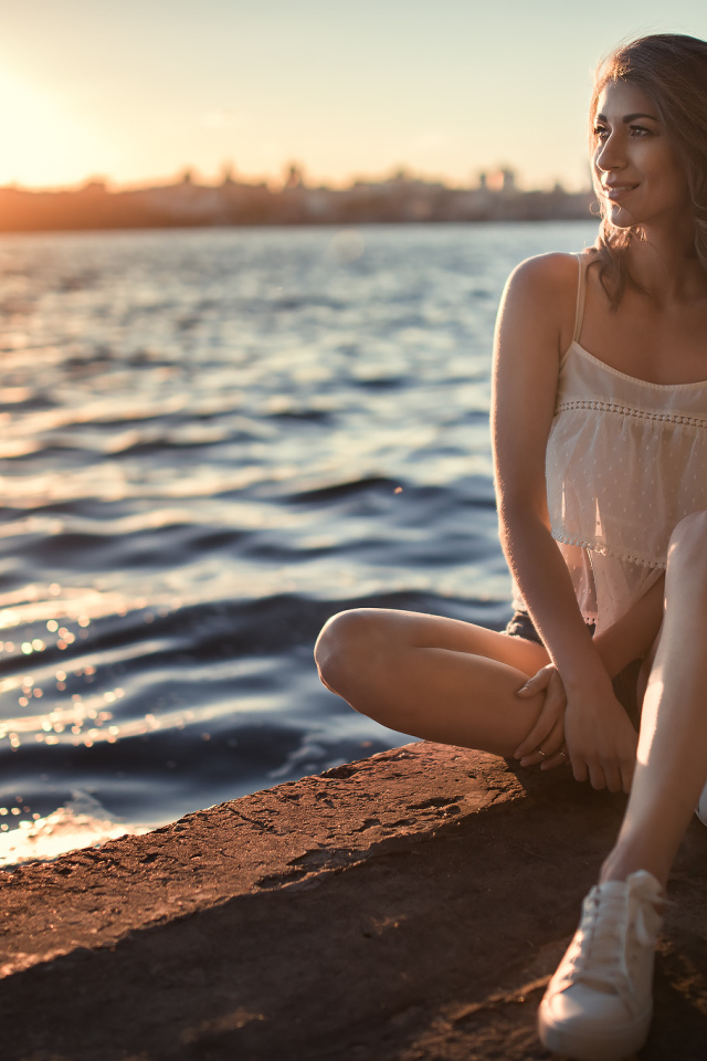 Красивая улыбающаяся девушка сидит в лучах солнца у реки