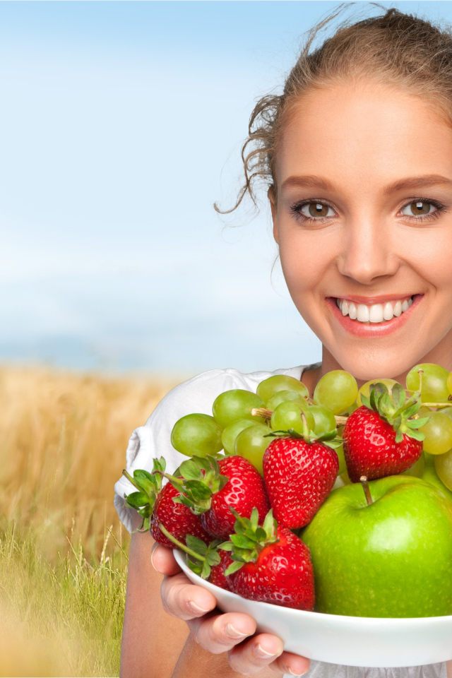 Улыбающаяся девушка шатенка с тарелкой с яблоками, виноградом, клубникой и грушей в руках