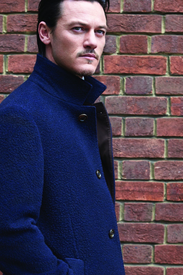 Актер Люк Эванс в синем плаще у кирпичной стены