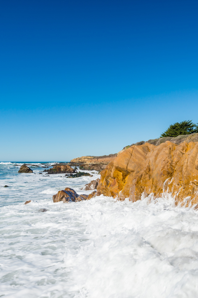 Белые волны бьются о скалы на берегу океана под голубым небом