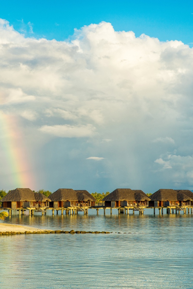 Bungalow in the water on a tropical beach under a cloudy sky with a rainbow