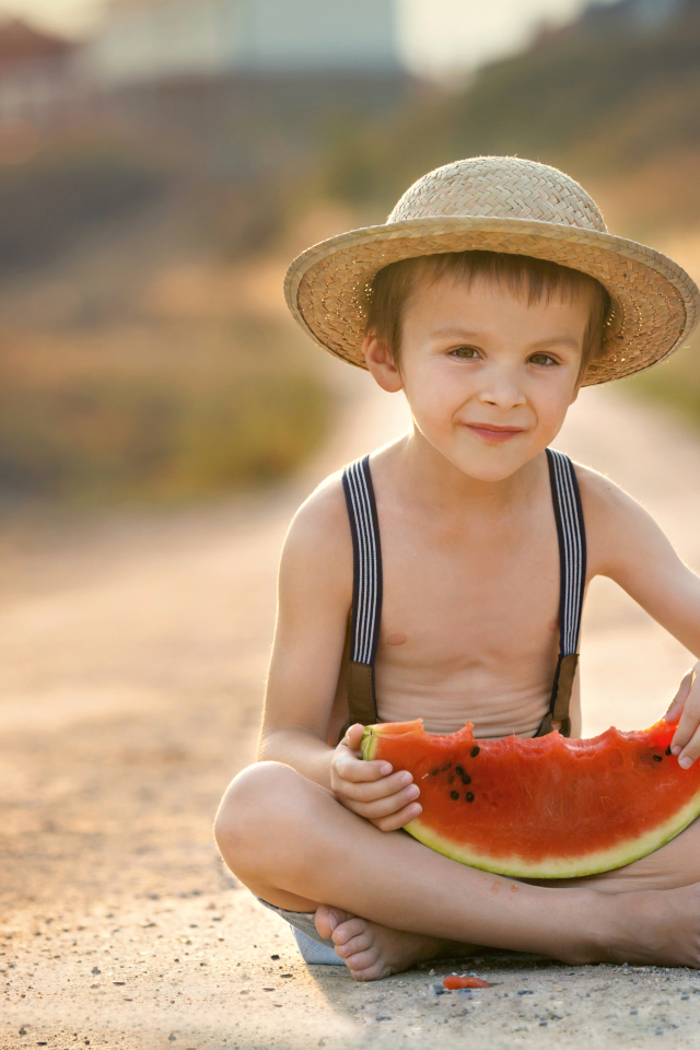 Маленький мальчик в шляпе сидит на дороге с долькой арбуза