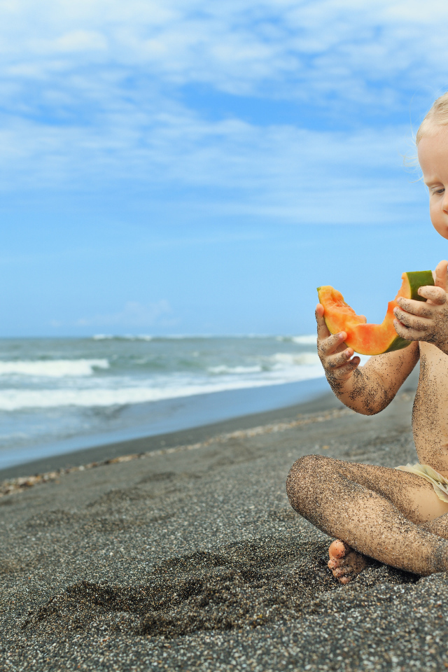 Маленькая девочка сидит на песке у моря с кусочком дыни