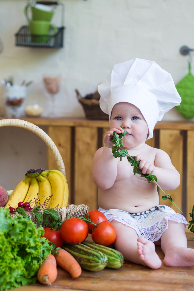 Маленький ребенок в шапке повара сидит на столе с овощами