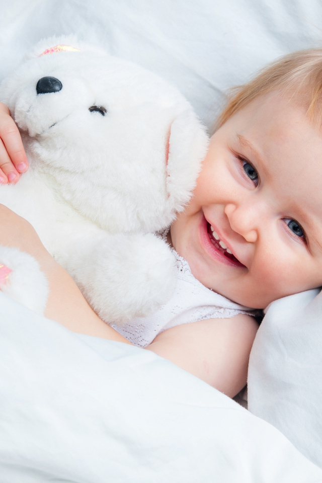 Улыбающийся ребенок лежит в кровати с белым плюшевым медвежонком