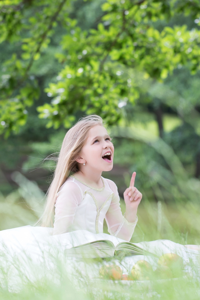 Красивая маленькая девочка в белом платье с книгой сидит на траве