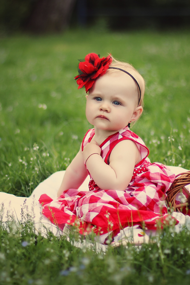 Красивая маленькая девочка с красным цветком на голове сидит на зеленой траве