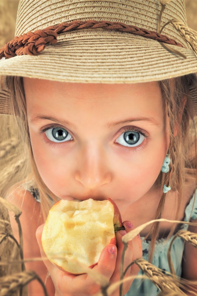 Маленькая голубоглазая девочка в шляпе ест яблоко