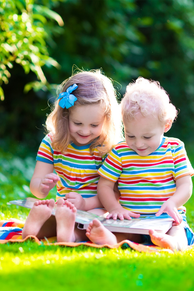Маленькие мальчик и девочка читают книгу сидя на зеленой траве