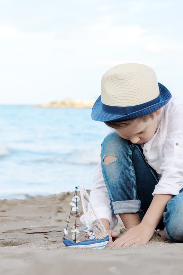 Маленький мальчик в шляпе играет с игрушечным корабликом на песке