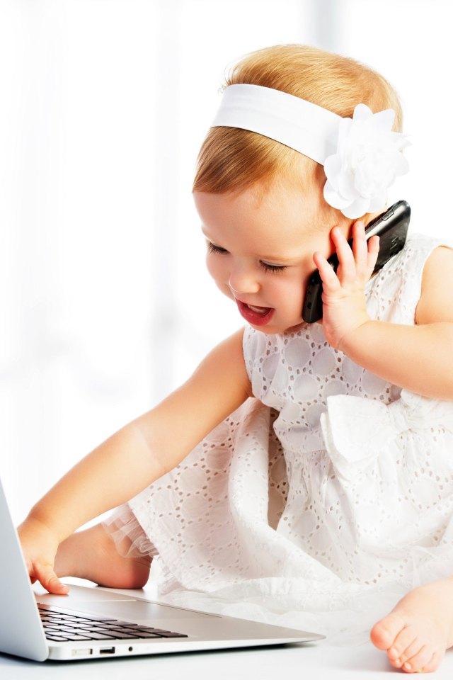 Маленькая девочка в белом платье сидит с телефоном у ноутбука
