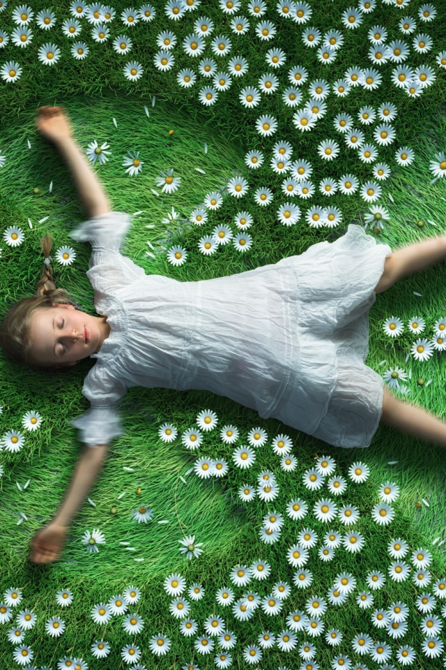 Маленькая девочка в белом платье лежит на зеленой траве с ромашками