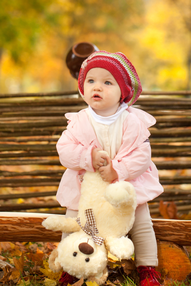 Маленькая девочка с плюшевым мишкой сидит на деревянной лавке осенью