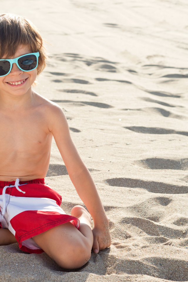 Маленький улыбающийся мальчик в очках сидит на песке