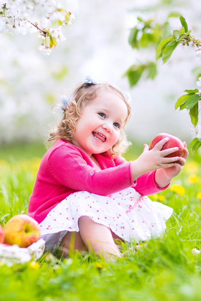 Маленькая улыбающаяся девочка сидит на зеленой траве с яблоками