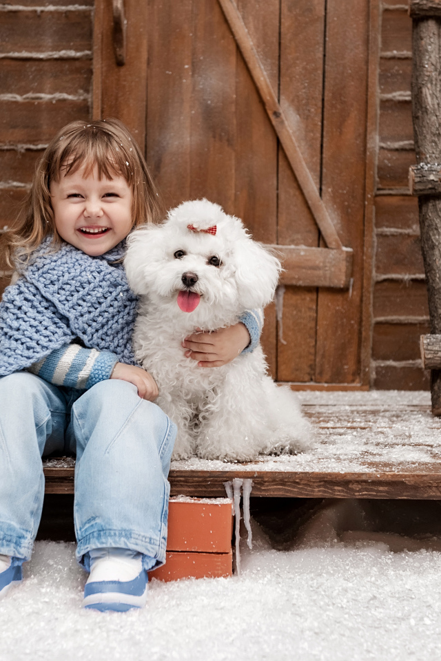 Маленькая улыбающаяся девочка сидит с белым пуделем на крыльце дома зимой