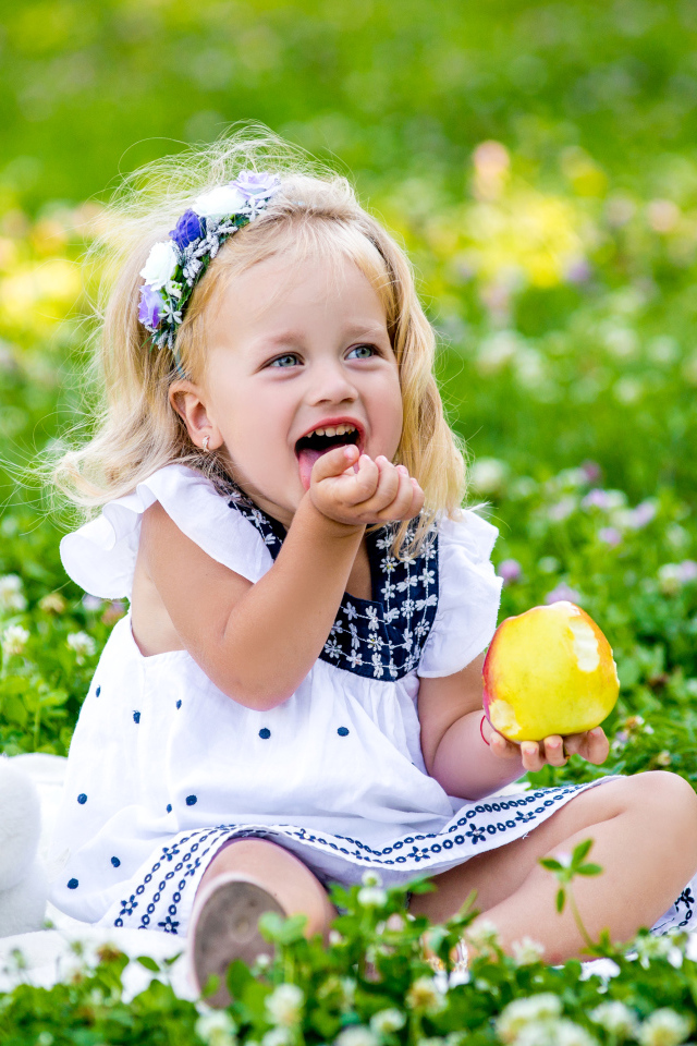 Маленькая улыбающаяся девочка с яблоком сидит на зеленой траве