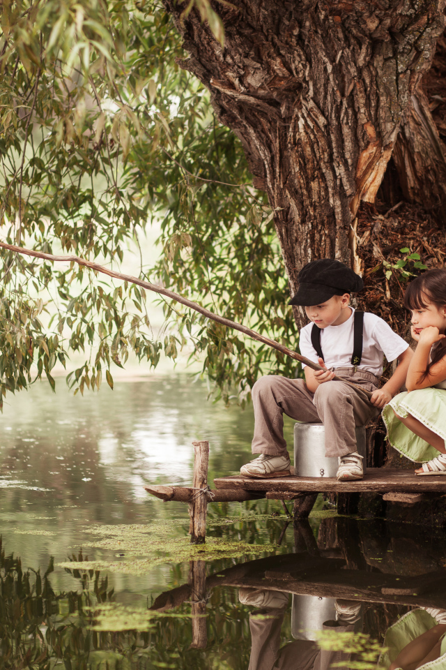 Две маленькие девочки и мальчик сидят на берегу пруда с удочкой