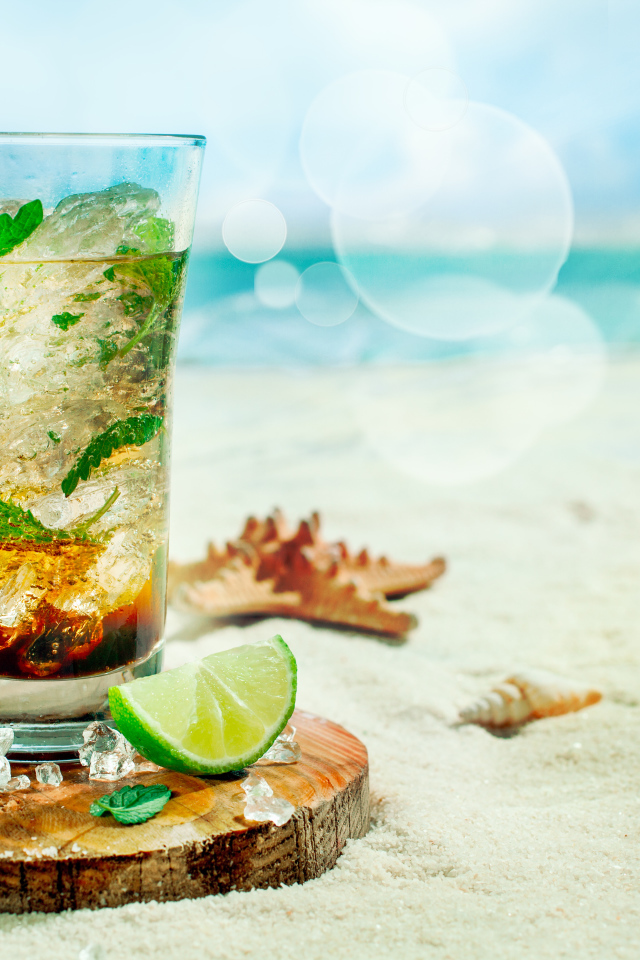 Коктейль в стакане с лаймом и кубиками льда стоит на песке на пляже