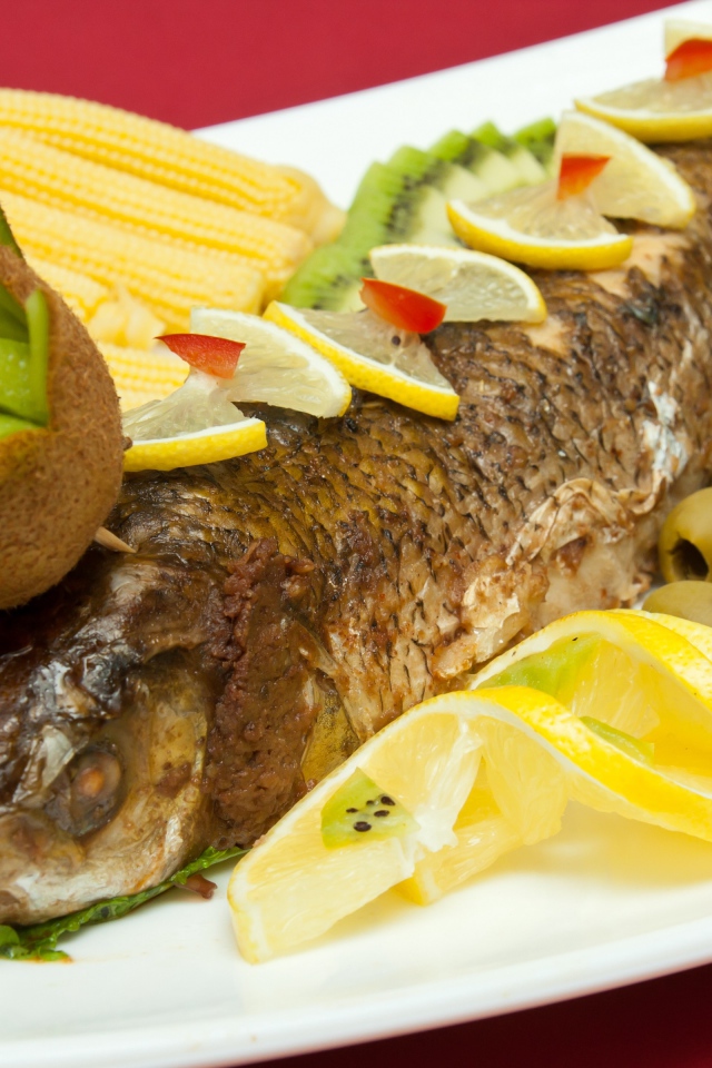 Запеченная рыба с оливками, лимоном, киви, кукурузой и листьями салата на белой тарелке