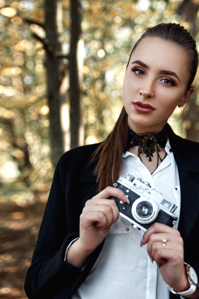 Красивая девушка с фотоаппаратом в руках стоит у дерева