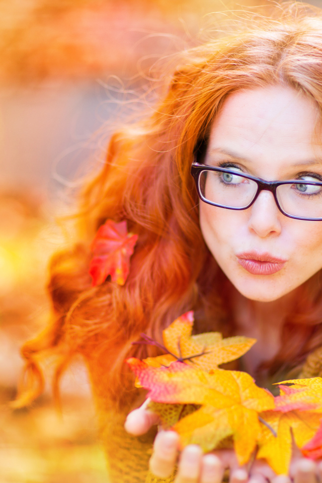 Рыжеволосая девушка в очках держит в руках опавшие листья 