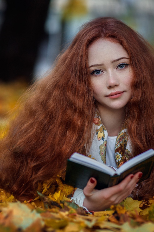 Рыжеволосая девушка с книгой лежит на опавших листьях осенью 