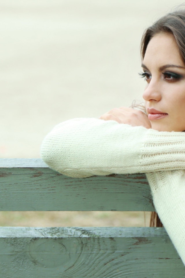 Задумчивая девушка брюнетка в белом свитере сидит на лавке