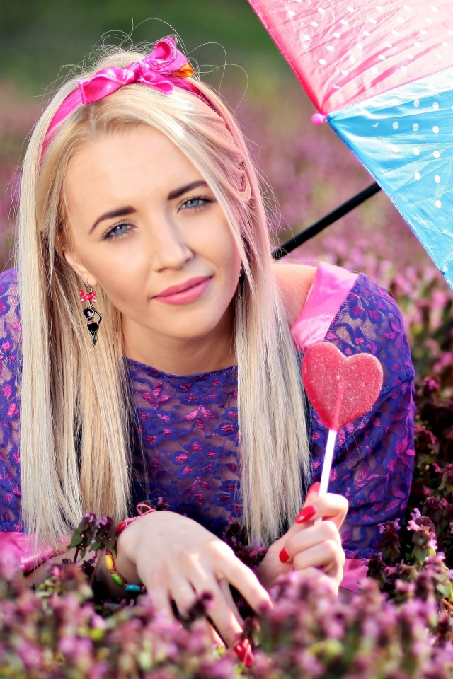 Молодая девушка блондинка лежит на поляне с сиреневыми цветами под разноцветным зонтом
