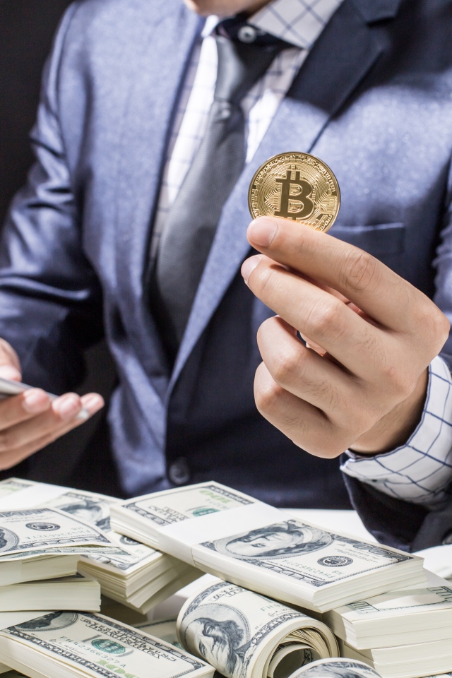 Мужчина с пачками долларов и монетой биткоин в руке