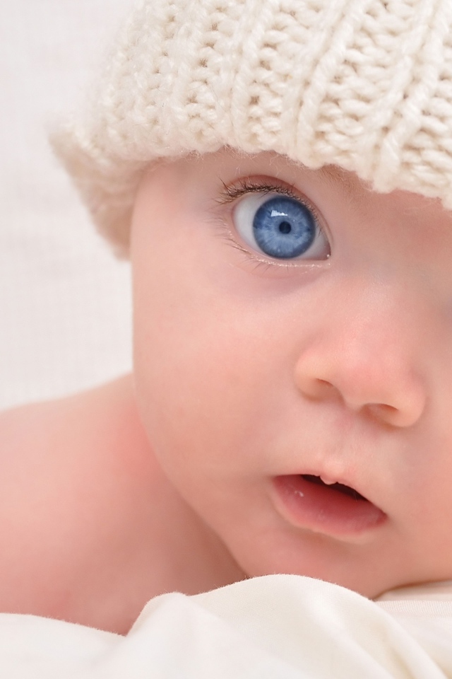 Голубоглазый малыш в белой вязаной шапке на сером фоне