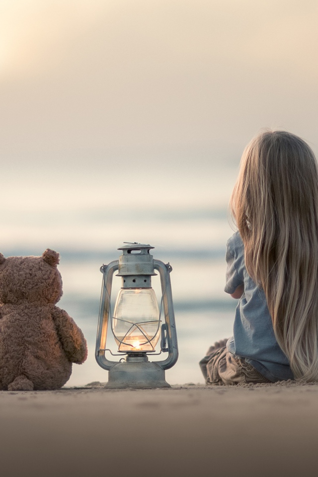 Маленькая девочка сидит на песке с плюшевым мишкой 