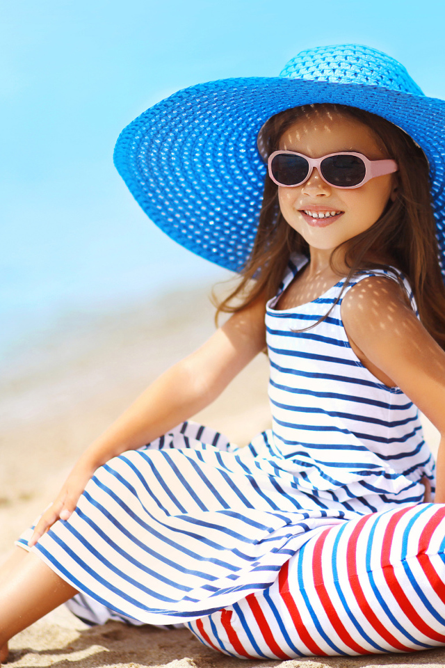 Маленькая девочка сидит на подушке на пляже в большой синей шляпе