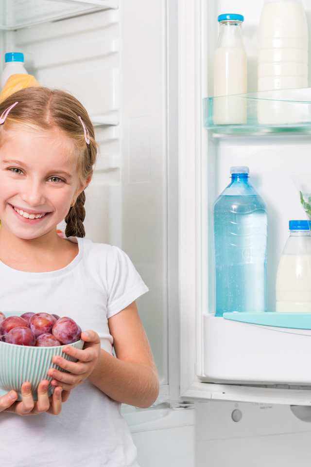 Маленькая девочка с миской со сливами в руках у холодильника 